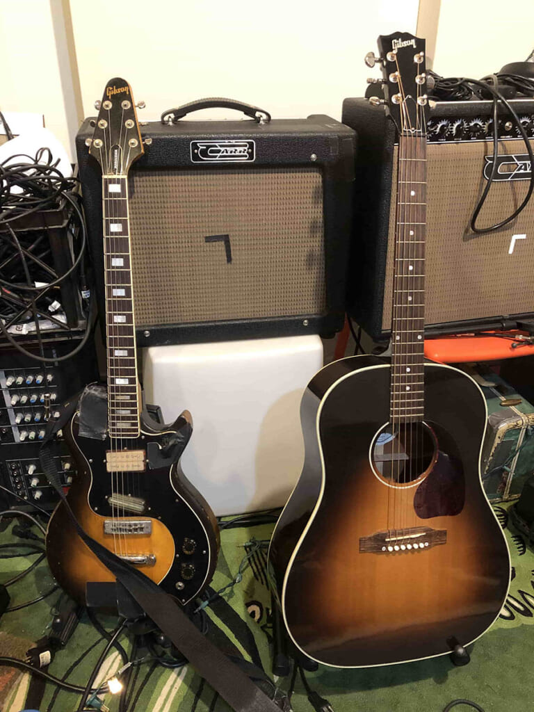 ギターは左が長年の愛器であるギブソン・マローダー・カスタム。1976〜77年のみ生産された貴重なモデルだ。右はギブソンJ-45で、エンドピンにシールドをつないで弾く姿も動画などで確認できるため、おそらく近年の現行品と思われる。また、ギターのうしろにあるのがレコーディングで使用したメイン・アンプのCarrのRambler（写真中央）とSlant 6V（写真右）。